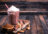 A tall glass of chocolate milkshake topped with whipped cream and a striped straw, surrounded by chocolate, cinnamon sticks, and almonds on a wooden surface.